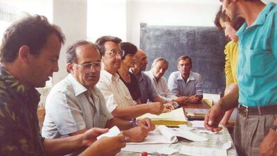 Election monitors seated around a table with papers, one standing; chalkboard in the background.