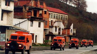 Convoy of orange armored vehicles driving along a war-torn street with damaged buildings.