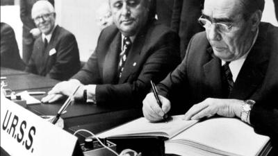 Black-and-white photograph of Leonid Brezhnev, General-Secretary of the Communist Party of the USSR, signing the Final Act at the Conference on Security and Co-operation in Europe, Helsinki, 1 August 1975. Brezhnev is focused on signing the document, with Turkey's Prime Minister Suleyman Demirel seated to his left, observing. A nameplate reading 'U.R.S.S.' is visible in the foreground, along with microphones and a folder.