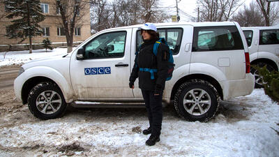 Person wearing winter gear stands beside a white SUV with OSCE logo on a snowy street, with buildings and trees in the background.