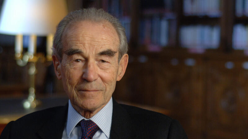 Elderly man in a suit sits in a dimly lit library, with bookshelves in the background.