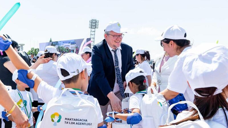 A man in a suit and cap interacts cheerfully with children wearing white caps and shirts, outdoors on a sunny day.