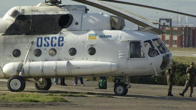 A white helicopter with the name of the OSCE and the Ukrainian flag standing on the tarmac of the Tbilisi military airport, with several soldiers standing nearby.