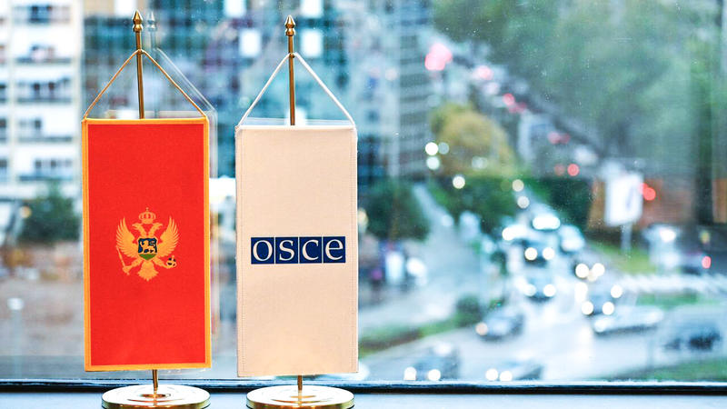 Two small desk banners—Montenegro’s red flag with a gold emblem and a white OSCE banner—stand on a window ledge with a blurred city street in the background.