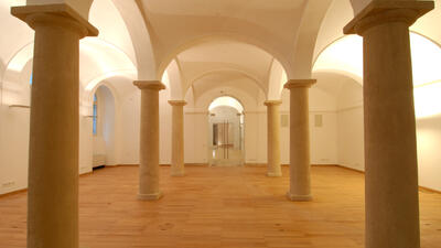 The image shows a spacious hall with classical architectural elements. The room features large, evenly spaced stone columns supporting a series of vaulted ceilings. The floor is made of warm-toned wood, contrasting with the white walls and ceilings. The lighting is soft and warm, enhancing the historical ambiance of the space.