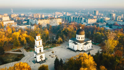 Cathedral and bell tower in a park with autumn trees, cityscape in the background.