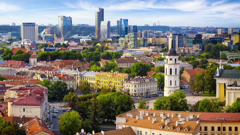 Skyline of Vilnius with colorful historic buildings, lush green trees, and modern skyscrapers under a blue sky.