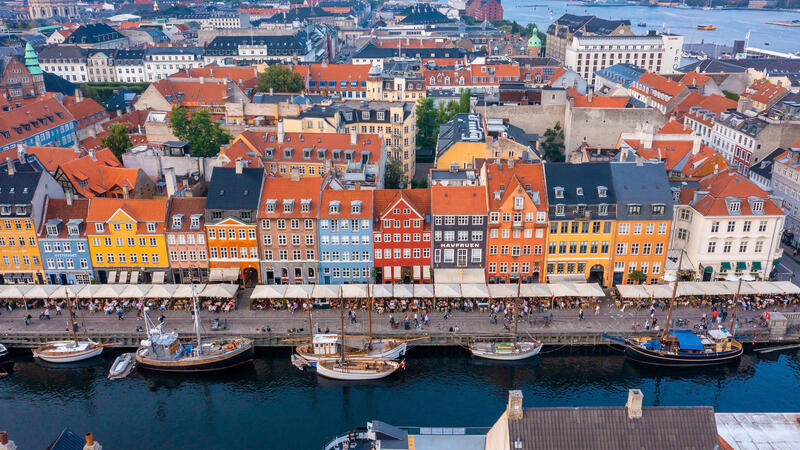 Colorful buildings line a canal in a European city, with boats moored along the water and a cityscape in the background.