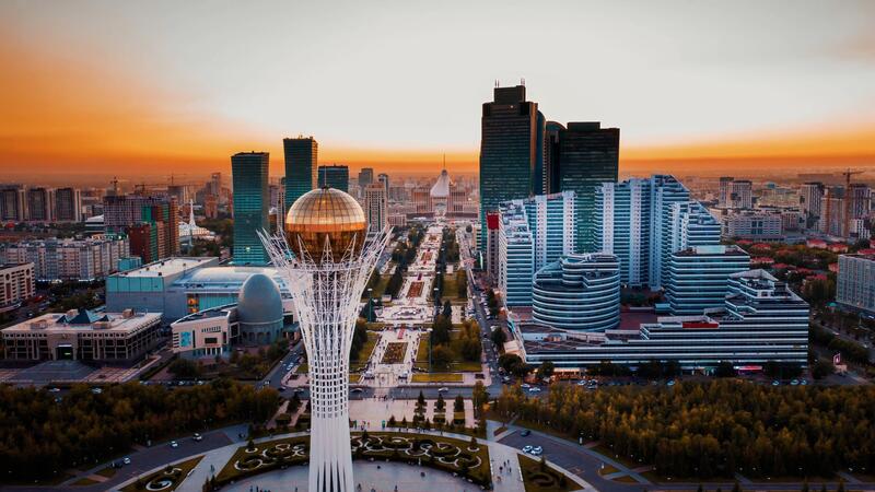 City skyline at sunset with a tall tower and modern buildings, featuring a golden sphere at the top.