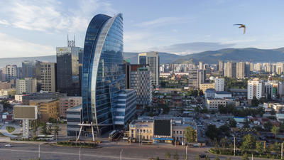 Modern cityscape with a distinctive blue glass skyscraper, surrounded by smaller buildings and mountains in the background.