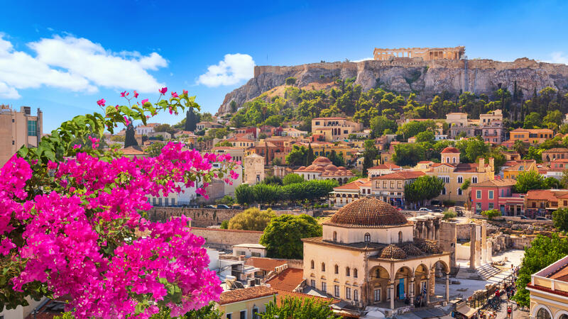 Athens cityscape with vibrant pink flowers, historic buildings, and the Acropolis perched on a hill under a clear blue sky.