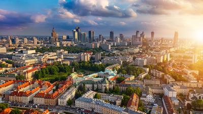 Aerial view of a cityscape at sunset, with skyscrapers, green parks, and historic buildings beneath a cloudy sky.