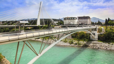 Cable-stayed bridge over turquoise river with city buildings and mountains in the background.
