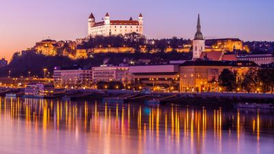 Bratislava Castle illuminated at sunset, reflecting on the Danube River, with a clear purple sky.