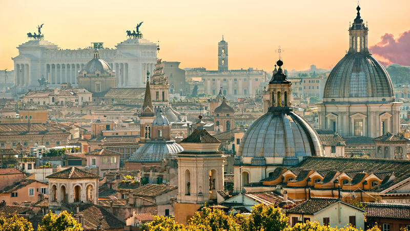 Skyline of Rome at sunset, featuring historic domes and buildings under a warm, orange sky.