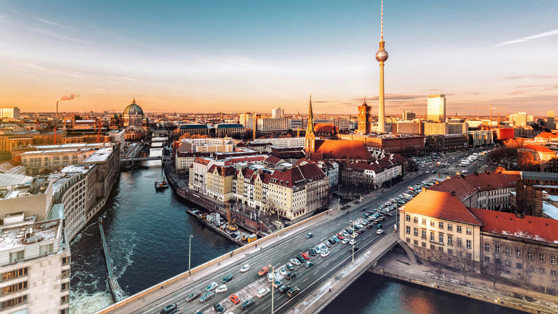 Aerial view of a cityscape at sunset with a river, bridges, and a tall TV tower in the distance.